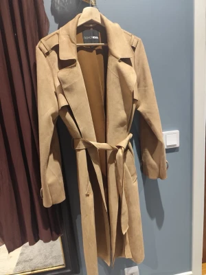 Beige kappa från Fashion Nova - Stilren beige kappa från Fashion Nova med klassisk krage, axelklaffar och knytskärp i midjan. Tillverkad i mjukt mockaimitationsmaterial och har långa ärmar samt slits baktill för extra rörelsefrihet. Perfekt för en trendig och avslappnad look. Storlek är M men passar L också. Använd 1 gång för test.