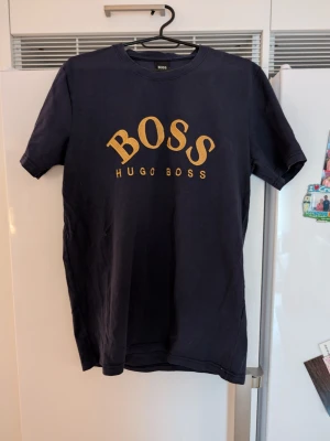 Svart Hugo Boss t-shirt med guldtryck - Snygg svart t-shirt från Hugo Boss med stort guldigt BOSS-tryck på bröstet. Klassisk rund halsringning och korta ärmar. Perfekt för dig som vill ha en stilren och trendig look med exklusiv känsla. Tillverkad i mjuk bomull för skön komfort. Mörkblå