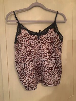 Leopardmönstrad linnetopp med spets - Säljer en leopardmönstrad linnetopp med tunna axelband och svarta spetsdetaljer vid urringningen. Toppen har en mjuk, glansig yta och en liten rosett framtill. Perfekt för dig som gillar djurmönster och vill ha något extra till outfiten.
