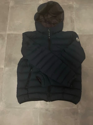 Marinblå  pufferjacka från Colmar - Snygg marinblå pufferjacka från Colmar med huva och dragkedja framtill. Jackan har ett broderat Colmar-märke på ärmen och två praktiska fickor med dragkedja. Perfekt för kalla dagar när du vill hålla värmen och ändå se stilren ut.