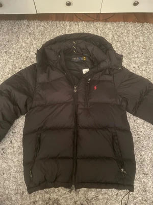 Svart dunjacka från Polo Ralph Lauren - Svart dunjacka från Polo Ralph Lauren med huva och dragkedja framtill. Jackan har två sidofickor med dragkedja och en bröstficka med dragkedja. Klassisk röd Polo-logga broderad på bröstet. Perfekt för kalla vinterdagar.