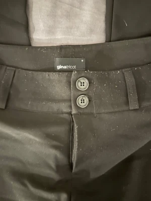Svarta kostymbyxor Gina Tricot - Snygga svarta kostymbyxor från Gina Tricot med bootcut, två knappar framtill och bälteshällor. Byxorna har två bakfickor med lock och knapp. Materialet är mjukt och har en lätt stretch. Köpta för 500 kr och säljer för 170 kr