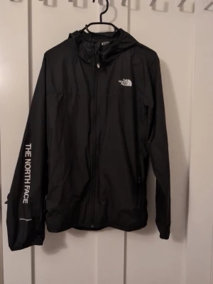 Svart vindjacka från The North Face - Snygg svart vindjacka från The North Face med huva och dragkedja framtill. Jackan är i storlek xl men samt barn storlek 14-16 år.Jackan har logga på bröstet och ärmen, samt praktiska fickor med dragkedja. Tillverkad i lätt och tåligt material som står emot vind. Perfekt för dig som vill ha en stilren och funktionell jacka.