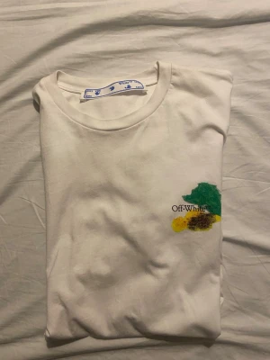 Vit Off-White t-shirt med färgglatt tryck - Vit t-shirt från Off-White med färgglada penseldrag och logga på bröstet. Stort konstnärligt tryck på ryggen i flera färger. Klassisk passform och rund halsringning. Perfekt för dig som gillar streetwear och unika detaljer.