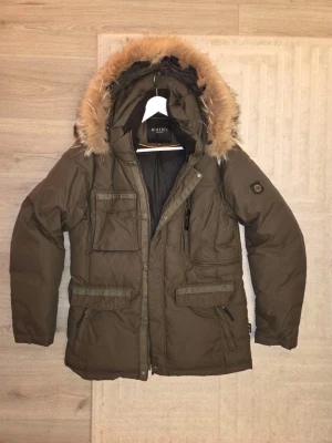 Olivgrön parkas från Hollies med äktapäls - Snygg olivgrön parkas från Hollies med stor huva och avtagbar päls. Jackan har flera praktiska fickor med lock och dragkedja, samt en klassisk rak passform. Perfekt för kalla dagar och ger en cool streetwear-look.