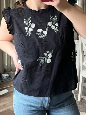 Svart blommig blus  - Säljer en svart blus med broderade blommor framtill. Blusen har volangdetaljer vid ärmarna och skurna detaljer framtill. 