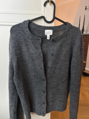 Mörkgrå kofta från H&M - Mysig mörkgrå kofta från H&M med rund halsringning och knäppning framtill. Tillverkad i mjukt stickat material och har långa ärmar. Perfekt att slänga över en t-shirt eller linne för en chill look.