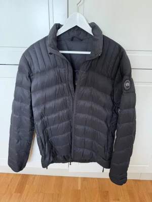 Svart dunjacka från Canada Goose - Snygg svart dunjacka från Canada Goose med quiltad design och hög krage. Jackan har dragkedja framtill, två sidofickor och en diskret logotyp på vänster ärm. Perfekt för kalla vinterdagar och riktigt varm tack vare dunfyllningen.  Väl använd därav priset -> säljes i befintligt skick