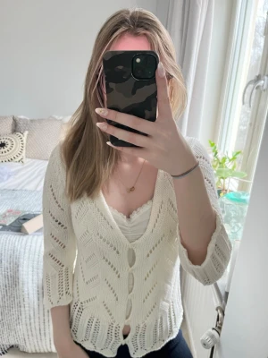 Vit virkad kofta med knappar - Snygg vit kofta med virkat mönster och trekvartsärmar. Djup V-ringning och dekorativa knappar framtill. Koftan har ett luftigt, stickat material och vågig kant nedtill. Perfekt att slänga över en topp för en chill look. Från Kappahl i storlek M