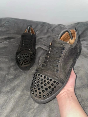 Christian Louboutin  - Säljer ett par svarta sneakers med coola svarta nitar på tån. Skorna har snörning, låg profil och är gjorda i mockaliknande material med brunt innerfoder. Perfekta för dig som vill sticka ut med din stil.