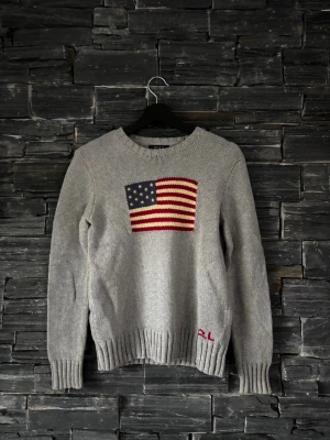 Grå stickad tröja med USA-flagga - Snygg grå stickad tröja från Ralph Lauren med en stor amerikansk flagga i rött, vitt och blått på bröstet. Tröjan har rund hals, ribbade muddar och RL-broderi vid nederkanten. Perfekt för dig som gillar klassisk college-stil.
