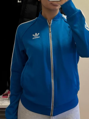 Blå Adidas zip hoodie med vita ränder - Snygg blå hoodie från Adidas med dragkedja och klassiska vita ränder längs ärmarna. Tröjan har ståkrage, två fickor och Adidas logga på bröstet. Perfekt för en sportig och avslappnad stil.