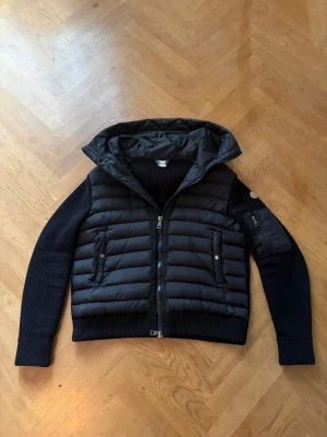 Svart pufferjacka med stickade ärmar - Svart pufferjacka från Moncler med quiltad kropp i polyester och stickade ärmar. Jackan har huva, dragkedja framtill, två fickor med knappar och en extra ficka på ärmen. Ribbad mudd vid ärmslut och nederkant för extra komfort.