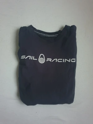 Marinblå Sail Racing t-shirt - Snygg marinblå t-shirt från Sail Racing med vit logga tryckt över bröstet. Klassisk rund hals och korta ärmar. Tillverkad i mjuk bomull som känns skön mot huden. Perfekt för dig som gillar stilren och sportig design.