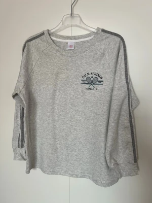 Grå sweatshirt Palm Springs - Grå sweatshirt med tryck 'Palm Springs Tennis Club' och två tennisrack på bröstet. Tröjan har rund hals, långa ärmar och mörkgrå ränder längs axlar och ärmar. Perfekt för en sportig och avslappnad stil.   Bredd (armhåla till armhåla): 50 cm • Längd (axel till nederkant): 62 cm • Ärmlängd (mätt från halsringningen): 61 cm Helt ny,aldrig använd,100%bomul 