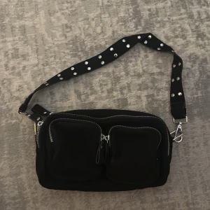 Svart axelväska med nitar från Gina Tricot  - Cool svart axelväska med två stora framfickor med dragkedjor, samt två stora fack. Axelremmen är dekorerad med silvriga nitar som ger en edgy vibe. Väskan är rektangulär och har en justerbar rem. Perfekt för dig som vill ha en praktisk men snygg accessoar.