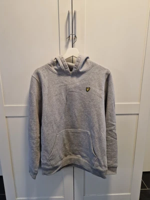 Grå hoodie från Lyle & Scott - Snygg grå hoodie från Lyle & Scott med klassisk gul logga på bröstet. Hoodien har huva, magficka och ribbade muddar vid ärmar och nederkant. Tillverkad i mjukt sweatshirtmaterial som är perfekt för en avslappnad stil.