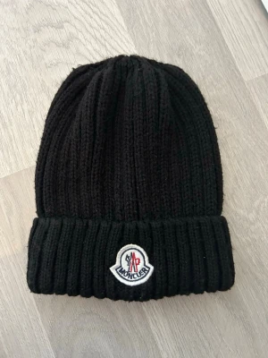 Svart ribbstickad mössa Moncler - Säljer en svart ribbstickad mössa från Moncler med uppvikt kant och klassisk Moncler-logga framtill. Mössan är gjord i tjockt, mjukt material som håller värmen och har en stilren look som passar till det mesta.