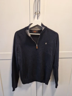 Mörkblå stickad tröja från Morris - Snygg mörkblå stickad tröja från Morris med halv dragkedja och liten logga broderad på bröstet. Tröjan har hög krage och ribbade muddar vid ärmslut och nederkant. Perfekt till lager på lager och enkel att matcha.