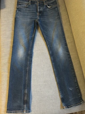 Blå jeans från Nudie Jeans - Snygga blå jeans från Nudie Jeans med rak passform. Jeansen har en fin tvätt och ljusare partier framtill samt rå kant vid benslut. Tillverkade i denim med kontrastsömmar och läderpatch bak i midjan. Modell grim tim 