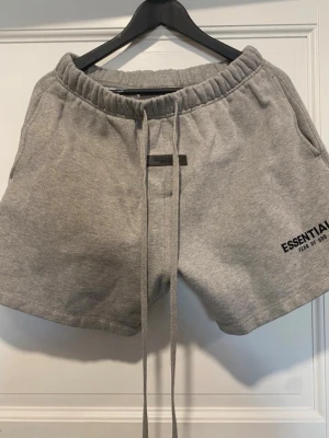 Grå shorts från Essentials - Gråa shorts från Essentials Fear of God med långa snören i midjan och elastisk midjeresår. Tillverkade i mjuk bomull, perfekt för chill dagar. Diskret logga tryckt på vänster ben. Två sidofickor och relaxed fit.
