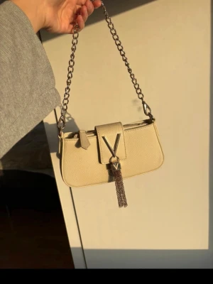 Valentino handväska - Snygg beige axelväska från Valentino med kedjerem i metall och V-logga framtill. Väskan är i skinnimitation med struktur och har dekorativt hänge i metallkedja. Perfekt storlek för det viktigaste och ger en lyxig känsla till din outfit. Enda defekten är att det som hämger från v:et har rostat och småfläckar inuti. Köpt på zalando!!!!Nypris 1200kr!!