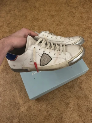 Vita sneakers från Philippe Model - Snygga vita sneakers från Philippe Model med grå detaljer och blå/röd häl. Skorna har en ikonisk sköldlogga på sidan och vita snören med en röd detalj. Tillverkade i läder med slitna effekter och grå sula. Perfekta för en trendig streetwear-look. Priset går att diskutera