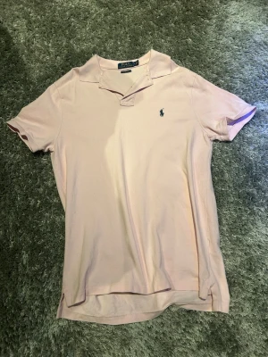 Ralph lauren piké  - Säljer en ljusrosa pikétröja från Ralph Lauren med klassisk krage och korta ärmar. Tröjan har den ikoniska broderade loggan i marinblått på bröstet. Tillverkad i mjuk bomull som känns skön mot huden. Perfekt för en clean och stilren look Tvär fet inför våren och sommarn.
