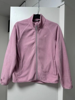 Rosa fleecekofta - Mysig rosa fleecekofta med hög krage och hel dragkedja framtill. Jackan har två sidofickor och är perfekt för lager-på-lager under kyliga dagar. Enkel och stilren design som passar till det mesta.