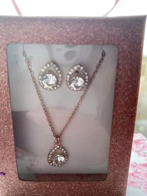Set med halsband och örhängen i guld - Säljer ett snyggt smyckeset med halsband och matchande örhängen i guldton. Både halsbandet och örhängena har droppformade hängen med gnistrande klara stenar och små kristaller runt om. Perfekt för dig som gillar elegant och glittrig stil.