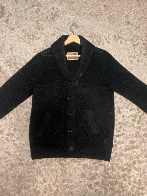 Svart kofta med sjalkrage från NOAH - Stilren svart kofta från NOAH med sjalkrage och knäppning framtill. Koftan har två fickor och är stickad i ett grovt material som ger en mysig känsla. Perfekt för lager på lager och enkel att matcha med olika outfits.