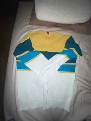 Flerfärgad stickad tröja med blockmönster - Säljer en stickad tröja med blockmönster i gult, vitt, blått och turkost. Tröjan har rund halsringning, långa ärmar och ribbade muddar. Materialet är mjukt och bekvämt, perfekt för dig som gillar färgglada plagg med modern känsla.