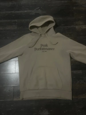 Beige hoodie från Peak Performance - Snygg beige hoodie från Peak Performance med tryck på bröstet och klassisk känguruficka. Hoodien har dragsko i huvan och ribbade muddar vid ärmslut och nederkant. Perfekt för en avslappnad och stilren look. Hör av er vid frågor!