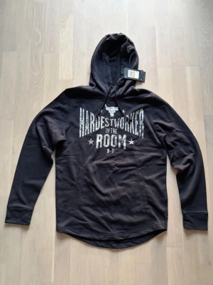Svart hoodie Under Armour  - Säljer en helt ny svart hoodie från Under Armour med trycket 'Hardest Worker In The Room' och tjur-logga på bröstet. Hoodien har huva, magficka och långa ärmar. 