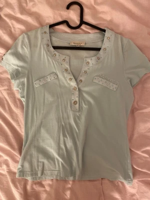 Ljusblå t-shirt med nitar - Snygg ljusblå t-shirt med korta ärmar och dekorativa nitar runt halsringningen och på bröstet. T-shirten har en v-ringad design med knappdetaljer och är gjord i mjuk bomull. Perfekt för en trendig och avslappnad look. Står i tröjan att storleken är M/L men jag som brukar ha Xs i tröja passar den bra på så skulle verkligen säga att tröjan är åt de mindre hållet.