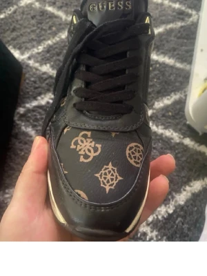 Svarta sneakers från Guess med logga - Snygga svarta sneakers från Guess med guldfärgade detaljer och klassiskt Guess-mönster i beige på ovandelen. Skon har svarta skosnören och en vit sula med Guess-logga baktill. Tillverkad i konstläder för en trendig look.