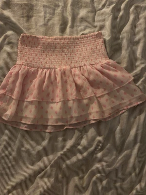 Rosa kortkjol med volang från Gina Tricot - Supersöt rosa kortkjol från Gina Tricot med smockad midja och dubbla volanger. Kjolen har ett ljust rosa tyg med små hjärtan i en mörkare rosa nyans. Perfekt för dig som gillar romantiska och lekfulla plagg.