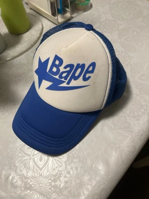 Blå och vit Bape truckerkeps - Säljer en snygg blå och vit truckerkeps från Bape med stort blått logotryck framtill. Kepsen har mesh på sidorna och bak för extra ventilation samt justerbar snapback. Perfekt för dig som gillar streetwear och vill sticka ut med en ikonisk accessoar.