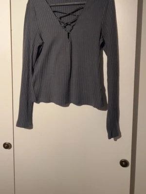 Mörkgrå ribbad topp med snörning - Snygg mörkgrå ribbad topp med lång ärm och v-ringning med snörningsdetalj framtill. Perfekt för dig som gillar en enkel men trendig stil. Materialet är mjukt och stretchigt för en bekväm passform.