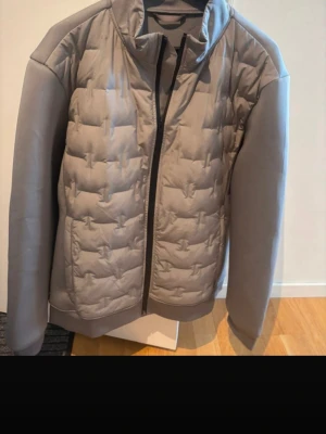 Grå pufferjacka från J.Lindeberg - Snygg grå pufferjacka från J.Lindeberg med quiltad front och släta ärmar. Jackan har hög krage, dragkedja framtill och diskret logga i nacken. Perfekt för kalla dagar och har en modern, stilren look.