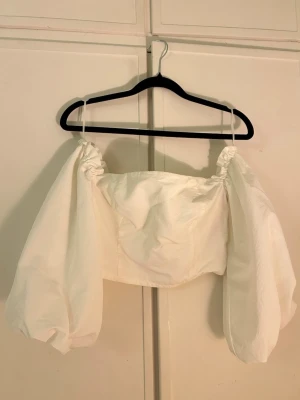 Vit offshoulder topp från Amisu - Trendig vit offshoulder topp från Amisu med puffiga, långa ärmar och smockad resår upptill. Toppen har en croppad passform och tunna axelband som ger en cool och luftig look. Perfekt att styla med höga jeans eller kjol.