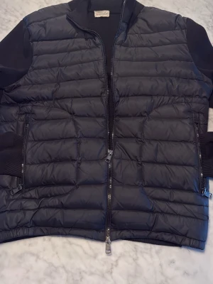 Svart Moncler cardigan  - Snygg svart dunväst med quiltad design och tvåvägsdragkedja framtill. Västen har ribbade muddar vid ärmarna och två fickor med dragkedja. Tillverkad i ett glansigt material och certifierad enligt DIST för spårbar dunfyllning.