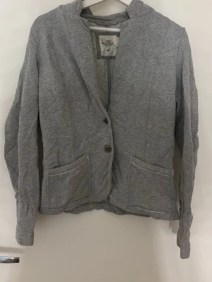 Grå kavaj i sweatshirtmaterial H&M - Mjuk grå kavaj från H&M i sweatshirtmaterial med två knappar framtill och två stora fickor. Rak passform och långa ärmar, perfekt för en avslappnad men ändå stilren look. Materialet är bomullsmix och färgen är ljusgrå.