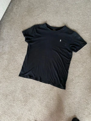 Svart t-shirt från Polo Ralph Lauren - En svart t-shirt från Polo Ralph Lauren med klassisk passform och korta ärmar. T-shirten har en liten broderad logotyp på bröstet och är tillverkad i mjuk bomull. Perfekt för dig som gillar stilrena basics med en touch av premium.