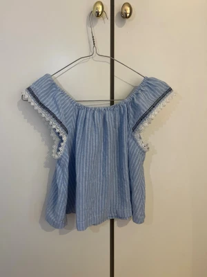 Blå blus från Mini malina - Supersöt blå- och vitrandig offshoulder blus från Maymalina. Blusen har volangiga ärmar med spetsdetaljer och en luftig passform. Perfekt för varma dagar och ger en fräsch sommarkänsla. Och knappt använd