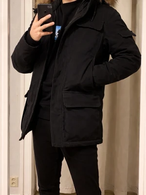 Svart vinterjacka  - Snygg svart parkas med huva och fuskpälsdetalj. Jackan har flera stora fickor framtill, dragkedja och vindslå. Perfekt för kalla dagar och har en klassisk, rak passform som funkar till det mesta.