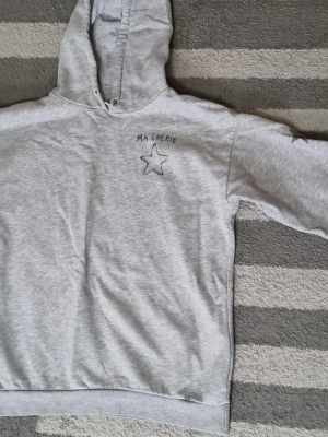 Grå hoodie med stjärntryck - Säljer en ljusgrå hoodie med huva och trycket 'MA CHERIE' samt en stjärna på bröstet. På ena ärmen finns flera stjärnor tryckta. Hoodien har ribbade muddar och är gjord i mjukt sweatshirtmaterial. Perfekt för en avslappnad och trendig look.
