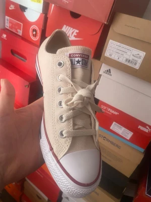 Beige Converse All Star låga sneakers - Säljer ett par klassiska Converse All Star sneakers i beige canvas med vita gummisulor och röda detaljer. Skorna har lågt skaft, vita snören och metallöljetter. Perfekta för en avslappnad och stilren look.