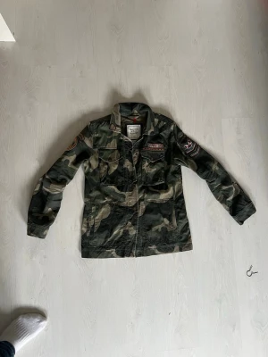 Camouflage vindjacka Abercrombie & Fitch - Säljer en gr sopgrön camouflage vindjacka från Abercrombie & Fitch med tryckknappar, fyra fickor framtill och coola patchdetaljer på bröstficka och ärm. Jackan har klassisk krage och är tillverkad i bomullsmix. Perfekt för digmesom gillar& militärstil och streetwear.