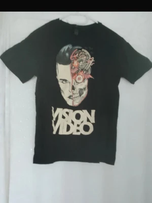 Svart Vision Video t-shirt - Cool svart t IC med tryck av ett  collaboration mellan människa och maskin på frams specificsidan samt texten 'VISION VIDEO' i vitt BF. T-shirten har rund hals och results korta ärmar. Perfekt för dig som gillar sopiga band och grafiska motiv.